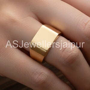 May include: A gold-colored ring with a rectangular top is worn on a finger. The ring has a smooth, polished surface. The text "ASJewellersjaipur" is printed on the finger. This ring is a piece of jewelry.