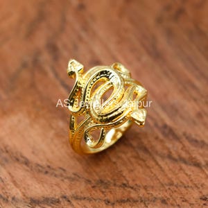 May include: A gold ring with an elaborate, swirling design. The ring has a detailed pattern with multiple loops and curves, creating a textured appearance. The ring is set against a warm, wooden background.