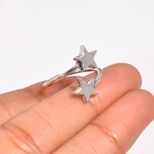 May include: A silver-toned open ring featuring two five-pointed star motifs at each end of the band. The stars are smooth and polished, positioned to overlap slightly, creating a bypass style design that wraps around the finger with a metallic finish.