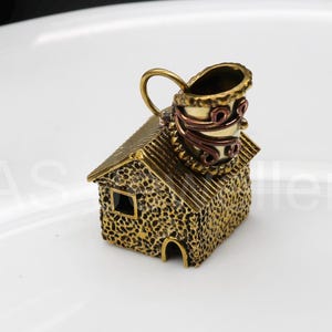 Può includere: Ciondolo a forma di casa in miniatura dettagliata con camino. La casa è strutturata con una finitura oro scuro, con una piccola finestra e una porta ad arco. Il camino è un mix di toni oro e rame, con volute decorative.
