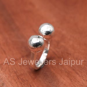Puede incluir: Un anillo de plata con dos adornos esféricos. El anillo tiene un diseño sencillo y moderno. El texto "AS Jewelers Jaipur" está impreso en la superficie.