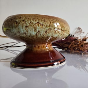May include: A ceramic pedestal bowl with a glossy finish. The bowl has a brown base and a wider top with a mottled brown and green glaze. The bowl is likely vintage and suitable for decorative purposes.
