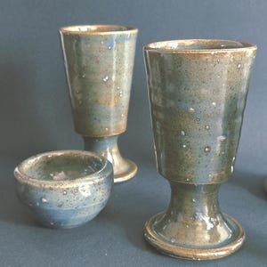 May include: A set of handmade ceramic drinkware. The set comprises two tall goblets and a small bowl, all with a speckled blue-green glaze and a brown rim. The pieces have a rustic, artisanal appearance, suitable for serving drinks.