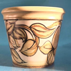 May include: A cream-coloured ceramic plant pot with a light brown rim. The pot features a leaf pattern in shades of brown and black. The design wraps around the exterior of the pot, adding a decorative touch.