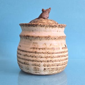 May include: A ceramic jar with a speckled cream and brown glaze featuring horizontal ridges along the body. The lid is topped with a small, sculpted brown mouse figure. The jar has a rounded shape and a wide opening, showcasing a rustic finish.