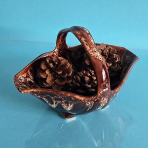 May include: A brown ceramic basket with a handle, filled with pine cones. The basket has a glossy finish with a speckled, two-tone design. The interior is divided into two sections, each holding several brown pine cones.