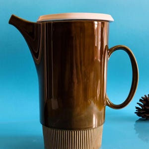 May include: A tall, brown ceramic coffee pot with a white lid and a handle. The base has a textured, vertical striped design. The spout is curved, and the pot is on a blue background. A pinecone is visible in the background.