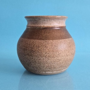 May include: A small, round ceramic vase with a speckled beige and tan finish. The vase features a distinct horizontal band of darker brown glaze around the upper middle section, a wide body, and a slightly flared rim with a smooth, rounded edge.