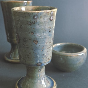 May include: Two ceramic goblets and one small ceramic bowl featuring a mottled blue and green glaze with dark brown speckles. The goblets have a tall, cylindrical body with a flared rim, a narrow stem, and a circular base, while the bowl is round.
