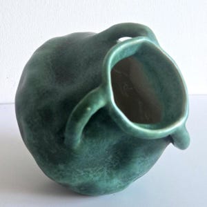 May include: A small, rounded ceramic vessel with a deep forest green glaze and a mottled, uneven surface texture. The piece features a wide, irregular opening and two small, curved handles attached to the sides of the main body.