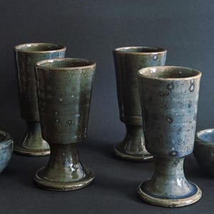May include: A set of handmade ceramic drinkware in blue and green hues. The collection features four stemmed goblets and two small bowls. The pottery has a speckled, textured finish, and a rustic aesthetic.