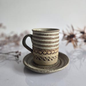 May include: A ceramic mug and saucer set. The mug has a handle and is beige with horizontal brown stripes and a decorative band of circles and triangles. The saucer is a matching beige colour. The set is on a white surface.