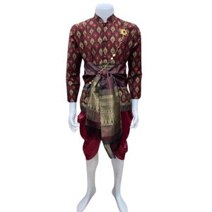 May include: A traditional Thai costume displayed on a mannequin. The outfit features a maroon and gold patterned long-sleeve shirt, matching pants, and a dark maroon sash. A gold brooch and decorative sash complete the ensemble. The costume is designed for formal occasions.