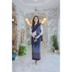 May include: A woman wearing a traditional navy blue outfit with a white blouse. The outfit includes a patterned skirt, a matching shawl, and a small patterned purse. The skirt and shawl feature intricate geometric designs in shades of blue, brown, and orange.