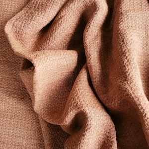 Può includere: Primo piano di un tessuto morbido e marrone con una superficie testurizzata. Il tessuto è piegato e drappeggiato, creando una sensazione di lusso e comfort.