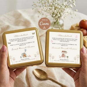 May include: Two gold-colored, square tins with printed announcements. The text reads "We're delighted to announce that our family has grown by two little feet and heartbeat." The tins are held by hands, with a spoon and sweets in the background.