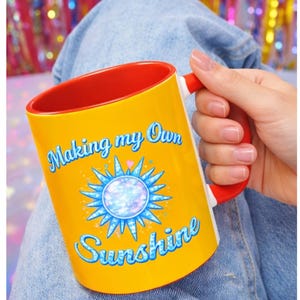 May include: Yellow ceramic mug with a red interior and handle. The mug features the text "Making my Own Sunshine" in a whimsical font, along with a blue and white sunburst design. The mug is held by a person wearing blue jeans.