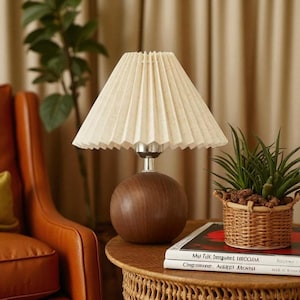 May include: A small table lamp featuring a cream pleated lampshade. The lamp has a spherical, wooden base and a silver stem. It sits on a small, round table, alongside a woven basket and books. The lamp's height is approximately 30 cm.