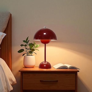 May include: A red, mid-century modern style table lamp with a rounded shade and a slender stem. The lamp is on a wooden nightstand next to a small potted plant and an open book. The lamp's height is approximately 12 inches.