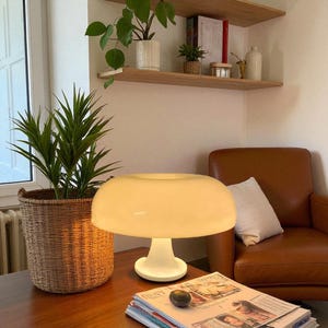 May include: A retro-style table lamp with a rounded, off-white shade and a white base. The lamp sits on a wooden table, next to a woven basket with a green plant. A brown leather armchair and a magazine are also visible.