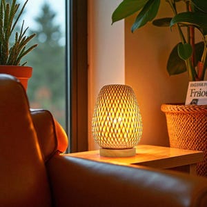 Puede incluir: Lámpara de mesa de bambú tejido con un brillo cálido. La lámpara tiene una base de madera y una pantalla de tejido abierto. La lámpara está sobre una mesa de madera junto a un sillón de cuero y una ventana con plantas. Altura: aprox. 20 cm.