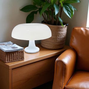 May include: A white, mushroom-shaped table lamp with a rounded shade and a pedestal base. The lamp sits on a wooden surface next to a woven basket and a potted plant. The lamp's design is minimalist and modern.