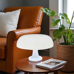 May include: A white mushroom-shaped table lamp with a rounded shade and a pedestal base. The lamp sits on a small, round, wooden table. A magazine is on the table. A brown leather armchair and a potted plant are in the background.