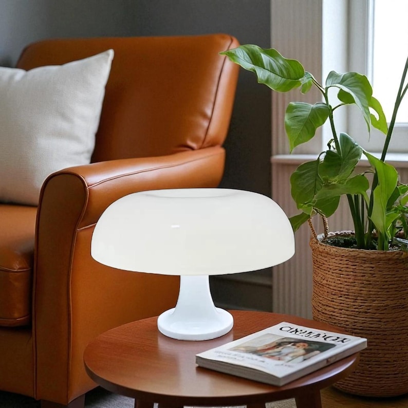 May include: A white mushroom-shaped table lamp with a rounded shade and a pedestal base. The lamp sits on a small, round, wooden table. A magazine is on the table. A brown leather armchair and a potted plant are in the background.