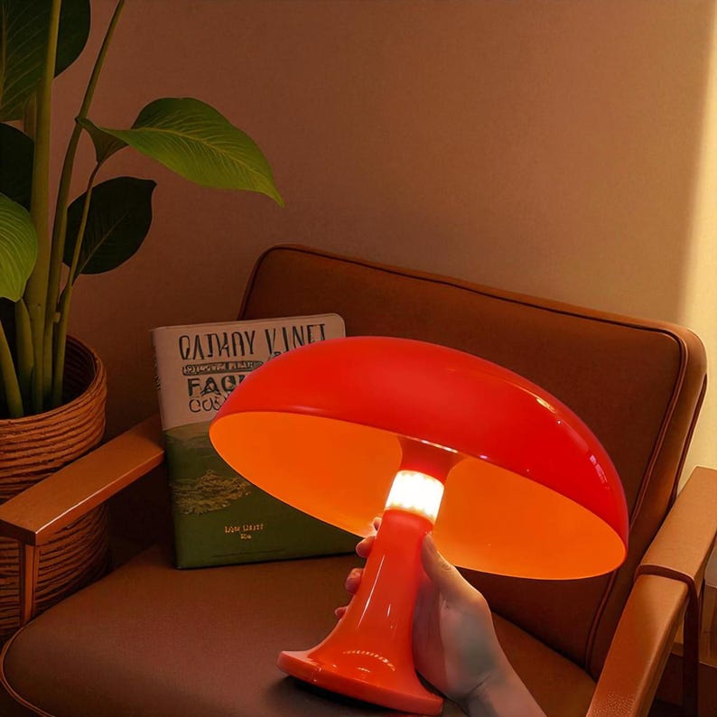 May include: A vibrant orange mushroom-shaped lamp with a bright orange interior. The lamp has a wide, rounded shade and a flared base. The lamp is held in a person's hand. A book with the title "CATHY VINET" is visible in the background.