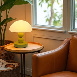 May include: A green mushroom-shaped table lamp with a round, frosted glass shade. The lamp sits on a wooden side table. The lamp's base and stem are a matching green colour. The lamp is next to a window and a brown leather armchair.