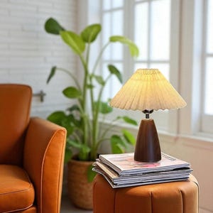 Pode incluir: Uma pequena lumin&aacute;ria de mesa com abajur plissado e base de madeira escura. A lumin&aacute;ria est&aacute; em cima de uma pilha de revistas em um pufe marrom. Uma planta em vaso e uma poltrona laranja est&atilde;o ao fundo.
