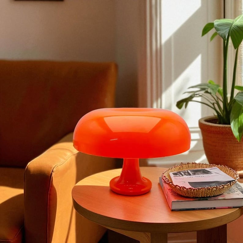 May include: A vibrant orange mushroom-shaped table lamp. The lamp has a rounded shade and a pedestal base. It sits on a round wooden table next to a gold-colored decorative bowl and a stack of books. The lamp is a retro design.