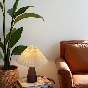 Pode incluir: Uma pequena lumin&aacute;ria de mesa com uma abajur plissado branco sujo e uma base de madeira escura. A lumin&aacute;ria est&aacute; em uma pilha de livros ao lado de uma planta em vaso e uma poltrona de couro marrom.