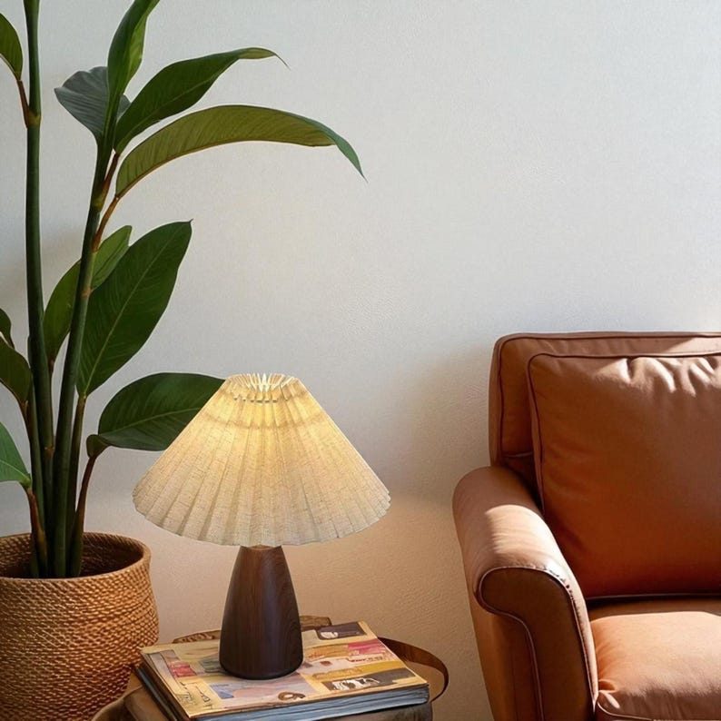 Pode incluir: Uma pequena lumin&aacute;ria de mesa com uma abajur plissado branco sujo e uma base de madeira escura. A lumin&aacute;ria est&aacute; em uma pilha de livros ao lado de uma planta em vaso e uma poltrona de couro marrom.