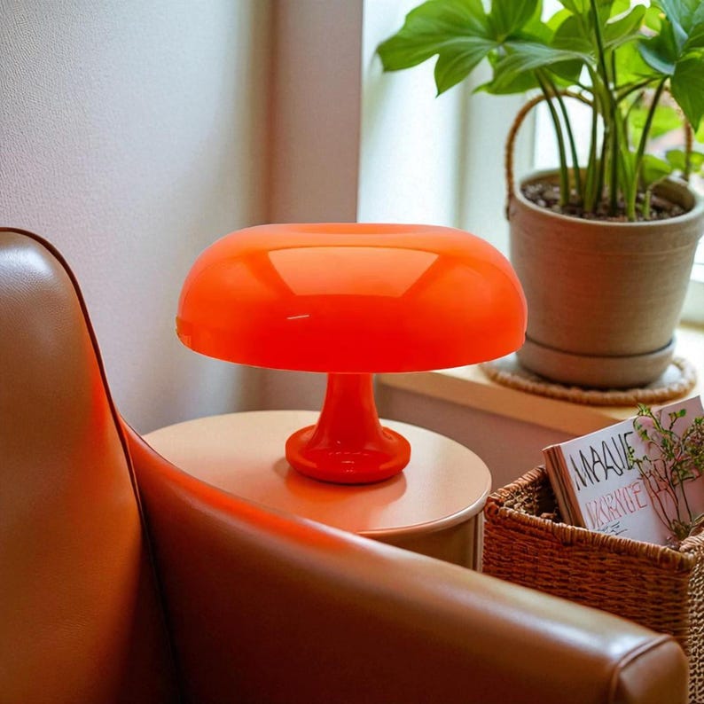 May include: A vibrant orange mushroom-shaped table lamp. The lamp has a rounded shade and a pedestal base. It sits on a small, round table next to a brown leather chair. A potted plant and a woven basket with a magazine are in the background.
