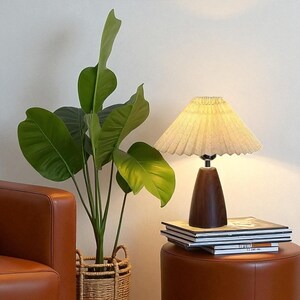 Pode incluir: Uma lumin&aacute;ria de mesa decorativa com abajur plissado e base de madeira escura. A lumin&aacute;ria est&aacute; sobre um pufe de couro marrom ao lado de uma pilha de livros. &Agrave; esquerda da lumin&aacute;ria, h&aacute; uma grande planta verde em uma cesta de vime.
