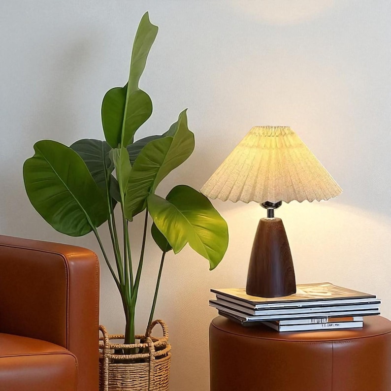 Pode incluir: Uma lumin&aacute;ria de mesa decorativa com abajur plissado e base de madeira escura. A lumin&aacute;ria est&aacute; sobre um pufe de couro marrom ao lado de uma pilha de livros. &Agrave; esquerda da lumin&aacute;ria, h&aacute; uma grande planta verde em uma cesta de vime.