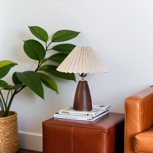 Pode incluir: Uma lumin&aacute;ria de mesa de madeira com uma sombra plissada bege est&aacute; em um pufe de couro marrom. A lumin&aacute;ria tem uma base de madeira escura e uma haste prateada. Uma pilha de revistas est&aacute; sob a lumin&aacute;ria. Uma planta em vaso com folhas verdes grandes est&aacute; ao fundo.