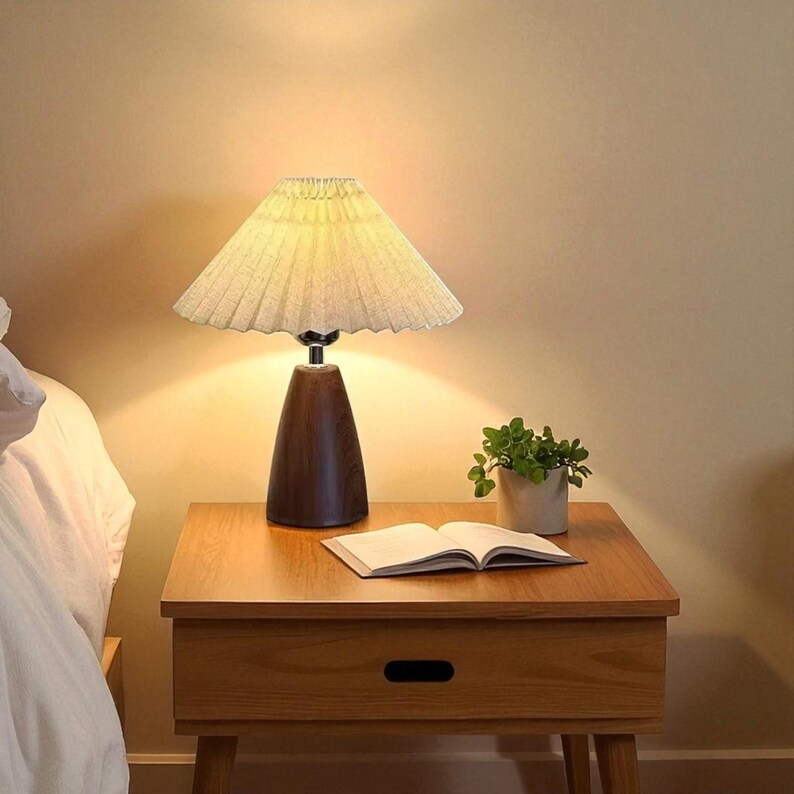 Pode incluir: Uma mesa de cabeceira de madeira com uma l&acirc;mpada, um livro e uma pequena planta em vaso. A l&acirc;mpada tem uma sombra plissada bege e uma base de madeira marrom escura. O livro aberto est&aacute; sobre a mesa, e a planta est&aacute; em um pequeno vaso claro.