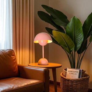 May include: A pink table lamp with a rounded shade and a slender stem sits on a small wooden table. The lamp's interior glows with a warm light. A brown leather armchair and a large potted plant with green leaves are in the background.