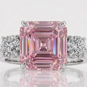 May include: A three-stone ring featuring a large, square-cut pink center stone held by four silver-toned prongs. The center stone is flanked by two smaller, round-cut clear stones on each side, all set on a polished silver-toned metal band.