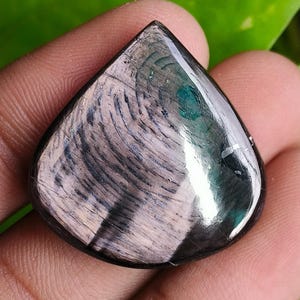 以下が含まれることがあります： 涙の形をした、研磨された宝石。石は、グレーと白の渦巻き模様で、ターコイズと黒のアクセントがあります。宝石は手で持たれており、背景には緑の葉があります。
