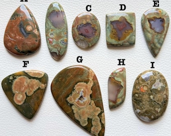 May include: Nine polished gemstones in various shapes, including teardrop, oval, and rectangular. The stones feature a mix of colors, such as brown, green, and purple, with unique patterns and textures. The gemstones are displayed on a light-colored surface.