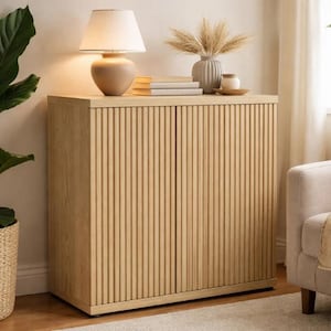 Peut inclure: Un buffet en bois clair avec des portes rainurées verticales. Une lampe avec un abat-jour et un socle beiges est posée sur le dessus, ainsi que des livres, un vase et une petite coupelle. Le buffet est à côté d'un fauteuil beige.