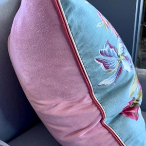 Puede incluir: Una almohada de terciopelo rosa con un estampado floral azul y ribete blanco. La almohada est&aacute; sentada en un sof&aacute; gris con tachuelas plateadas.