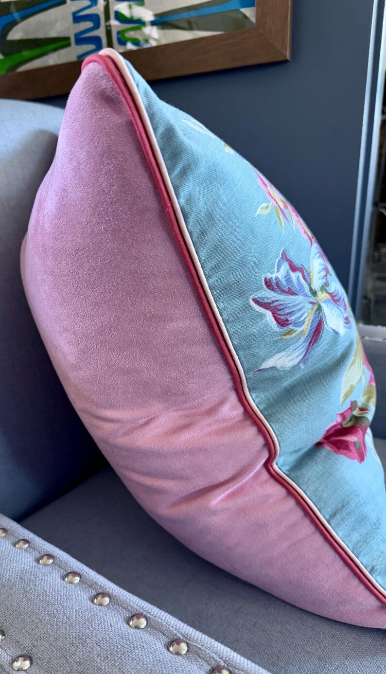 Puede incluir: Una almohada de terciopelo rosa con un estampado floral azul y ribete blanco. La almohada est&aacute; sentada en un sof&aacute; gris con tachuelas plateadas.