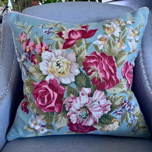 Puede incluir: Una almohada floral azul con una variedad de flores rosas, blancas y rojas sobre un fondo azul claro. La almohada est&aacute; sentada en una silla gris con tachuelas plateadas.