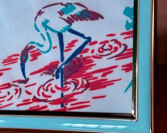 May include: A light blue picture frame with a silver trim, featuring a white fabric with a red and blue design of two birds standing in water.