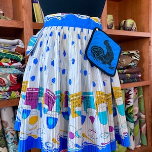 Puede incluir: Un delantal de algodón blanco con ribete azul y un agarrador de olla de gallo azul. El delantal tiene un estampado de copas de vino y botellas con el texto "Wines, Nosellie, Port, Sherry, Dive, Chill".