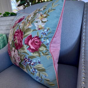 Puede incluir: Una almohada floral con flores rosas y blancas sobre un fondo azul claro. La almohada tiene un borde rosa y est&aacute; sentada en un sof&aacute; azul claro.